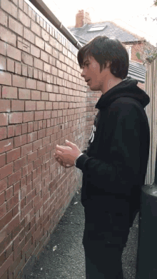 a young man wearing a black hoodie with the letter n on it stands in front of a brick wall