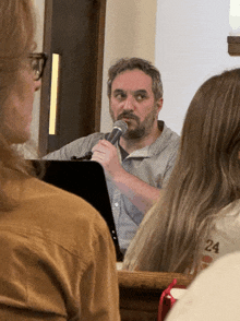 a man speaking into a microphone in front of a group of people with one wearing a 24 shirt