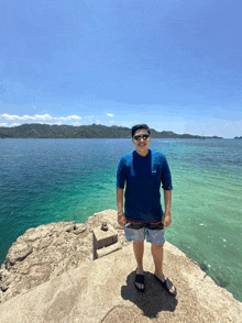 a man wearing sunglasses and a blue shirt stands on a rock overlooking the ocean