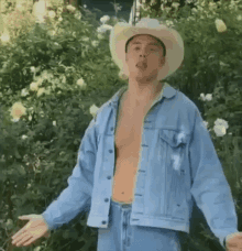 a shirtless man wearing a cowboy hat and a denim jacket is standing in front of flowers .