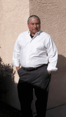 a man in a white shirt and black pants stands in front of a wall with his hands in his pockets