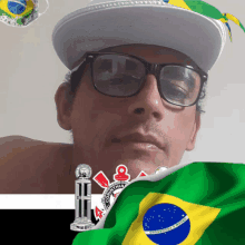 a man wearing glasses and a hat stands in front of a brazilian flag and a corinthians logo