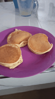 three pancakes on a purple plate with one bite taken out of one of them