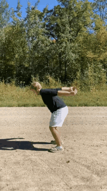 a man in a black shirt and white shorts is squatting on a dirt field in front of trees .