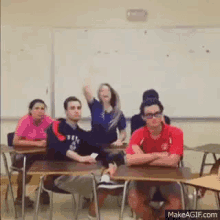 a group of students are sitting at desks in a classroom with a girl raising her hand in the air .