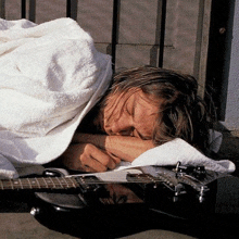 a man wrapped in a white towel sleeping next to a black guitar