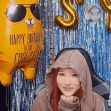 a woman wearing a hoodie is sitting in front of a birthday balloon that says happy birthday to one cool cat .