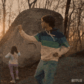 a man in a striped hoodie stands in front of a large rock with a netflix logo on the bottom right