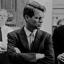 a man in a suit and tie is sitting with his arms crossed in a black and white photo .