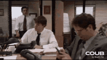 two men are sitting at a desk in an office looking at their cell phones .