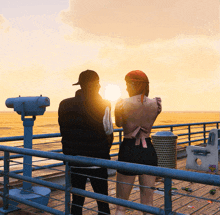 a man and a woman are standing on a pier watching the sunset