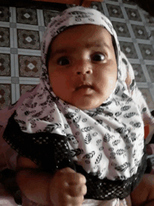 a baby wearing a white and black hijab looks at the camera