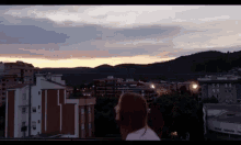 a woman stands on a balcony overlooking a city at sunset with a building that says ' allianz ' on it