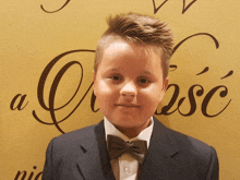 a young boy wearing a suit and bow tie stands in front of a wall that says a czasc