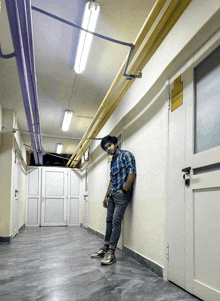 a man in a plaid shirt leans against a wall in a hallway with a yellow sign on the door that says exit