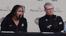 a man and a woman are sitting at a table with microphones in front of a wall that says basketball