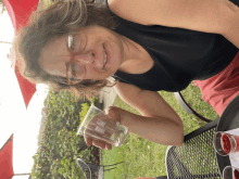 a woman wearing glasses sits at a table holding a clear plastic cup