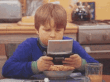 a young boy sitting at a table with a bowl of cereal and a glass of orange juice playing a video game