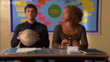 a man and a woman are sitting at a desk in front of a bulletin board that says bbc one