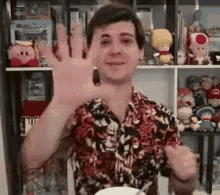 a man in a hawaiian shirt is waving his hand in front of a shelf filled with stuffed animals .