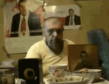 a man with a beard is sitting at a table with a bowl of food and a picture of a man on the wall .