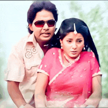 a man wearing sunglasses stands next to a woman in a pink saree