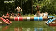 a man and a woman are jumping over a wooden beam in the water while a crowd watches in the background