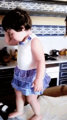 a little girl in a blue and white dress is standing in a kitchen and covering her face with her hands