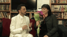 a man in a white suit is giving a woman a bouquet of flowers in front of a bookshelf that has sonic on it