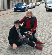 two men sitting on a cobblestone street with a blue car in the background that says peugeot