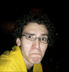 a man with curly hair wearing glasses and a yellow hoodie makes a funny face