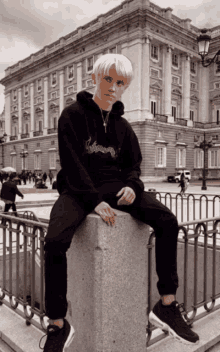 a young man in a black hoodie is sitting on a stone pillar in front of a large building