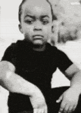 a black and white photo of a young boy sitting down with his arms crossed and making a funny face .