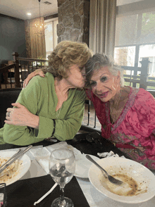 a woman kisses another woman on the cheek while they sit at a table