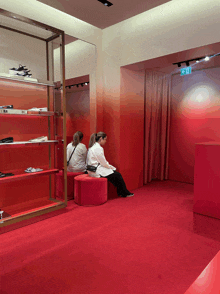 two women sit on red ottomans in a store with a red exit sign on the wall