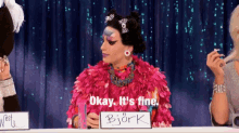 a drag queen is sitting at a table with a sign that says okay it 's fine bjork