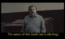 a man with a beard is standing in front of a wall with the words " the name of this trash can is ideology " above him