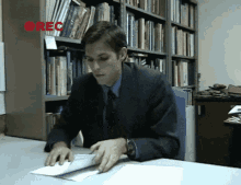 a man in a suit sits at a desk in front of a bookshelf with the word rec on it