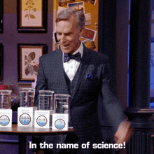 a man in a suit and bow tie stands in front of a table full of measuring cups and boxes that say in the name of science