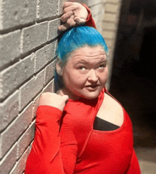 a woman with blue hair is leaning against a brick wall wearing a red top .