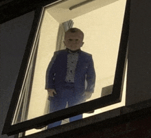 a little boy in a suit and bow tie stands in a window