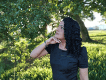 a woman in a black dress is standing in a field with her hand on her chin