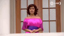 a woman in a pink off the shoulder dress stands on a balcony with a tvgo logo behind her