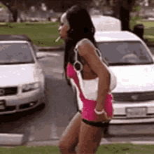 a woman in a pink dress is smoking a cigarette while standing in front of a white car .