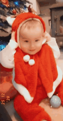 a baby dressed in a santa suit is sitting in front of a christmas tree ..