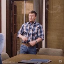 a man in a plaid shirt and jeans is standing in front of a table in a conference room .