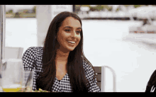 a woman in a plaid shirt is smiling while sitting at a table in front of a body of water