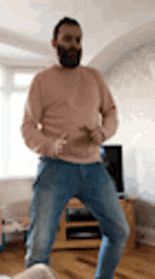 a man with a beard is dancing in a living room while wearing a pink sweater and jeans .