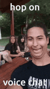 a man in a black shirt is taking a selfie with two women sitting at a picnic table behind him .