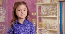 a little girl in a blue shirt is standing in front of a shelf filled with trophies and crowns .